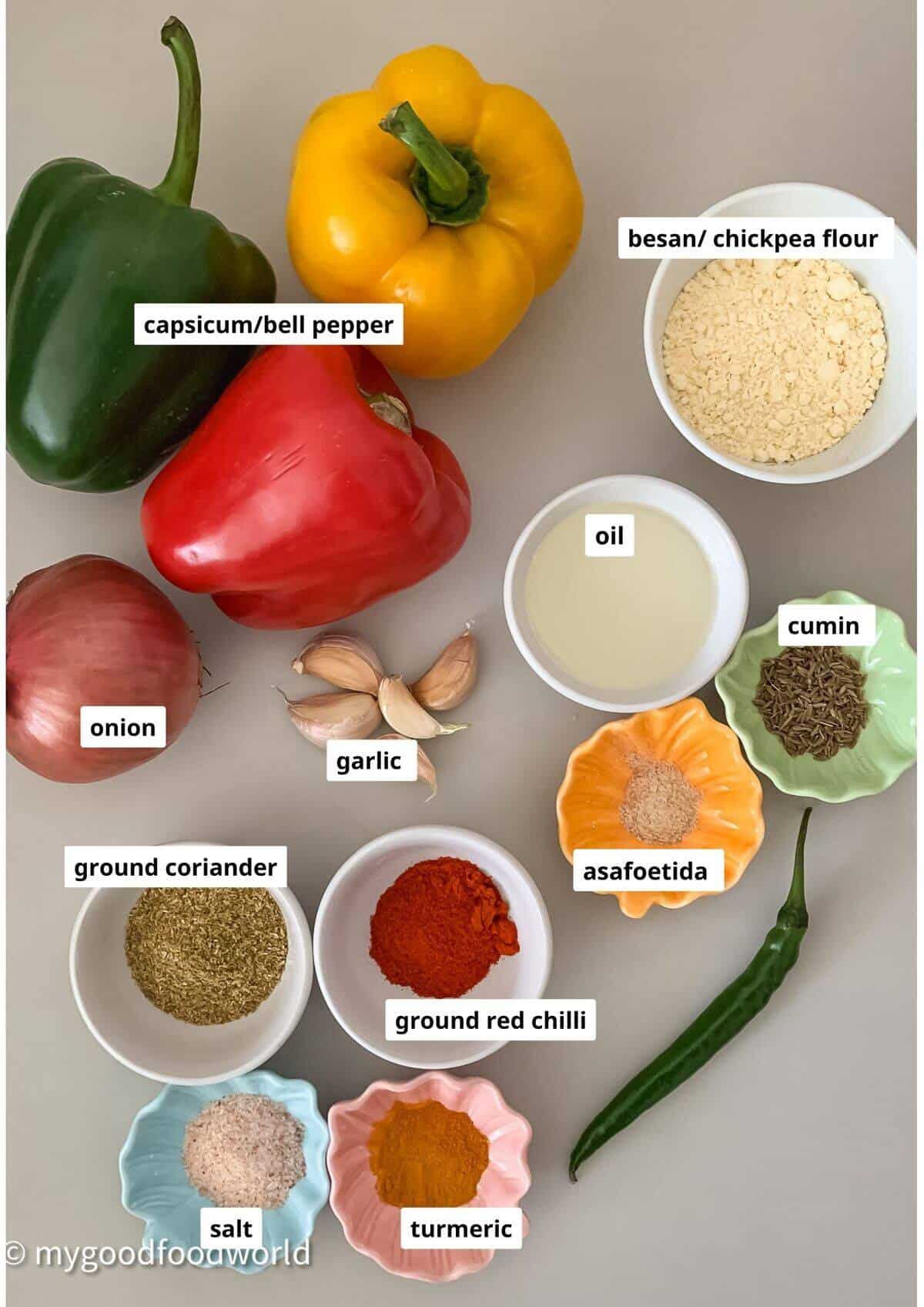 Ingredients for besan capsicum recipe such as tri colored bell peppers, gram flour, cumin seeds, chilli powder, and aromatics are placed on a light coloured background for a photo.