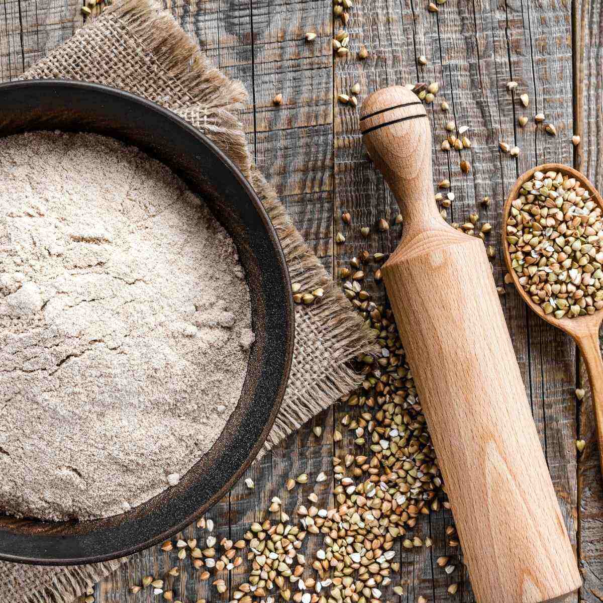 Buckwheat flour placed in a bowl with some groats spilled around it and a rolling pin next to the bowl.
