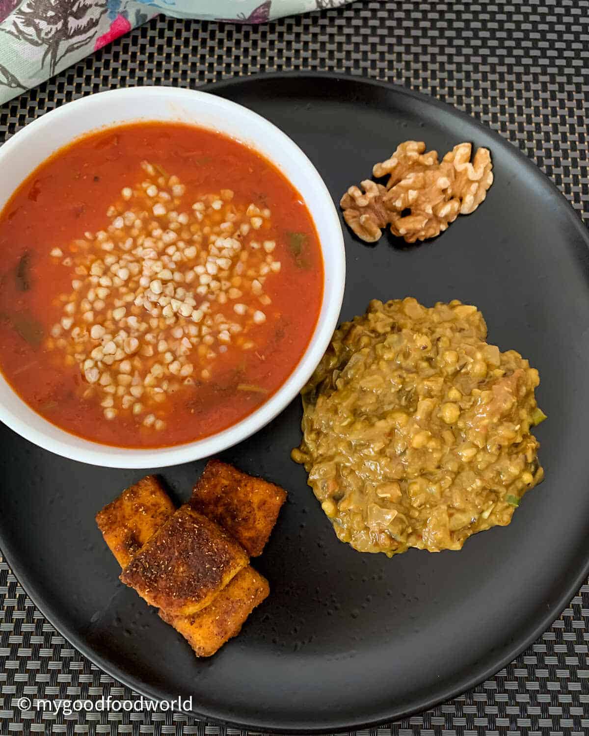 Buckwheat with tomato stew, beans curry, walnuts and pan fried paneer served on a black plat and placed on a black mat.