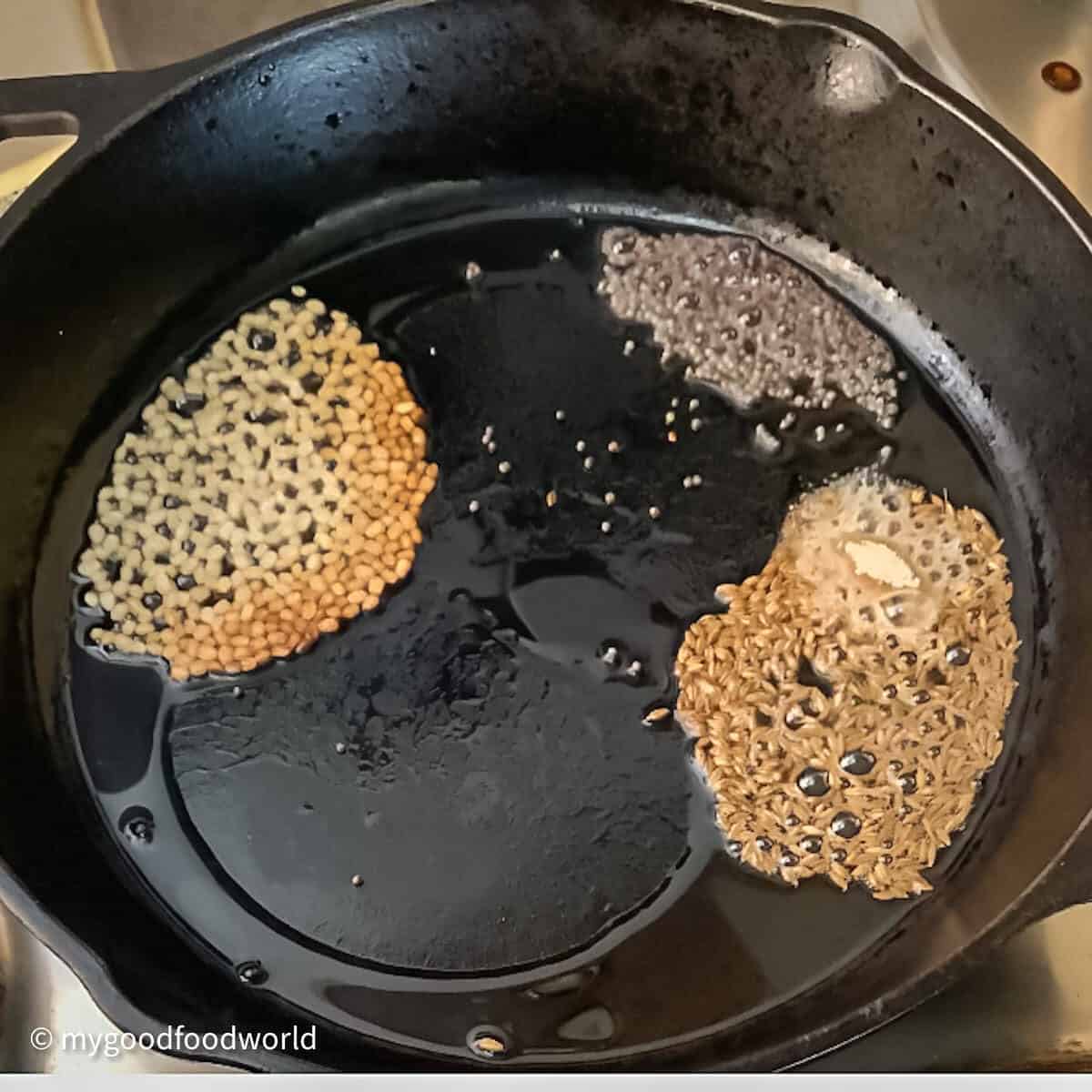 Lentils, cumin and mustard seeds frying in oil in a black colored cast iron pan.