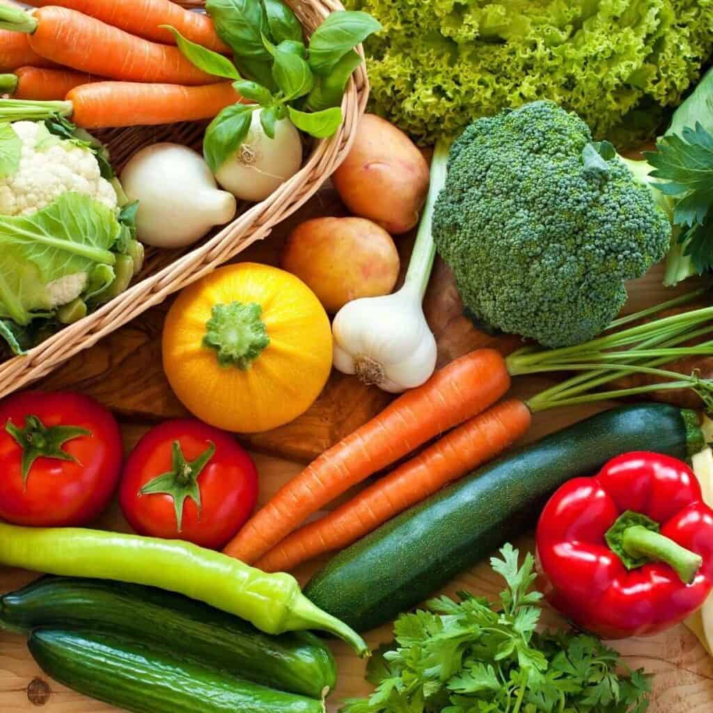 A collection of colorful vegetables such as carrots, broccoli, peppers, zucchini, cauliflower, and garlic in a basket and on a wooden board.