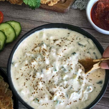 Cucumber raita recipe made and served in a round bowl with sliced cucumbers and pickle on the side.