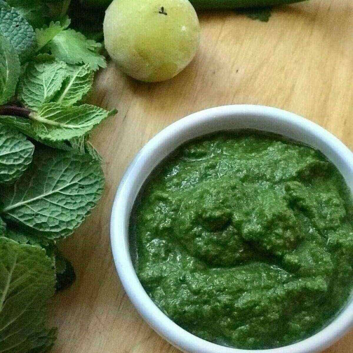 Cilantro mint chutney made with gooseberry, fresh coriander, mint and green chillies served in a round white bowl and placed on a table with fresh greens on the side.