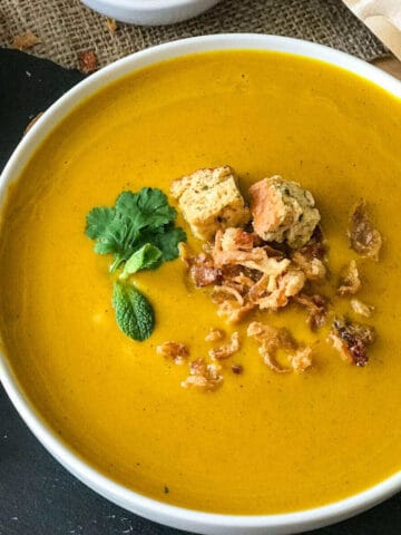Bright yellow vegan sweet potato soup served in a white round bowl with garnish of fresh mint leaves, croutons and fried onions.