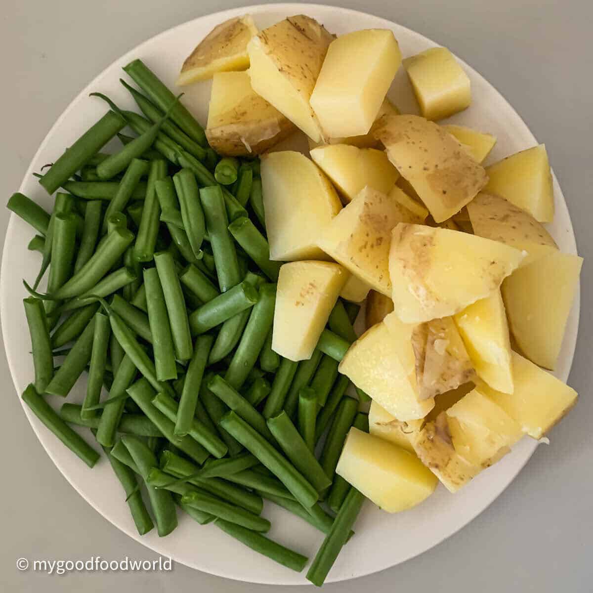 Chopped potatoes and french beans placed in a white plate on a light grey background.