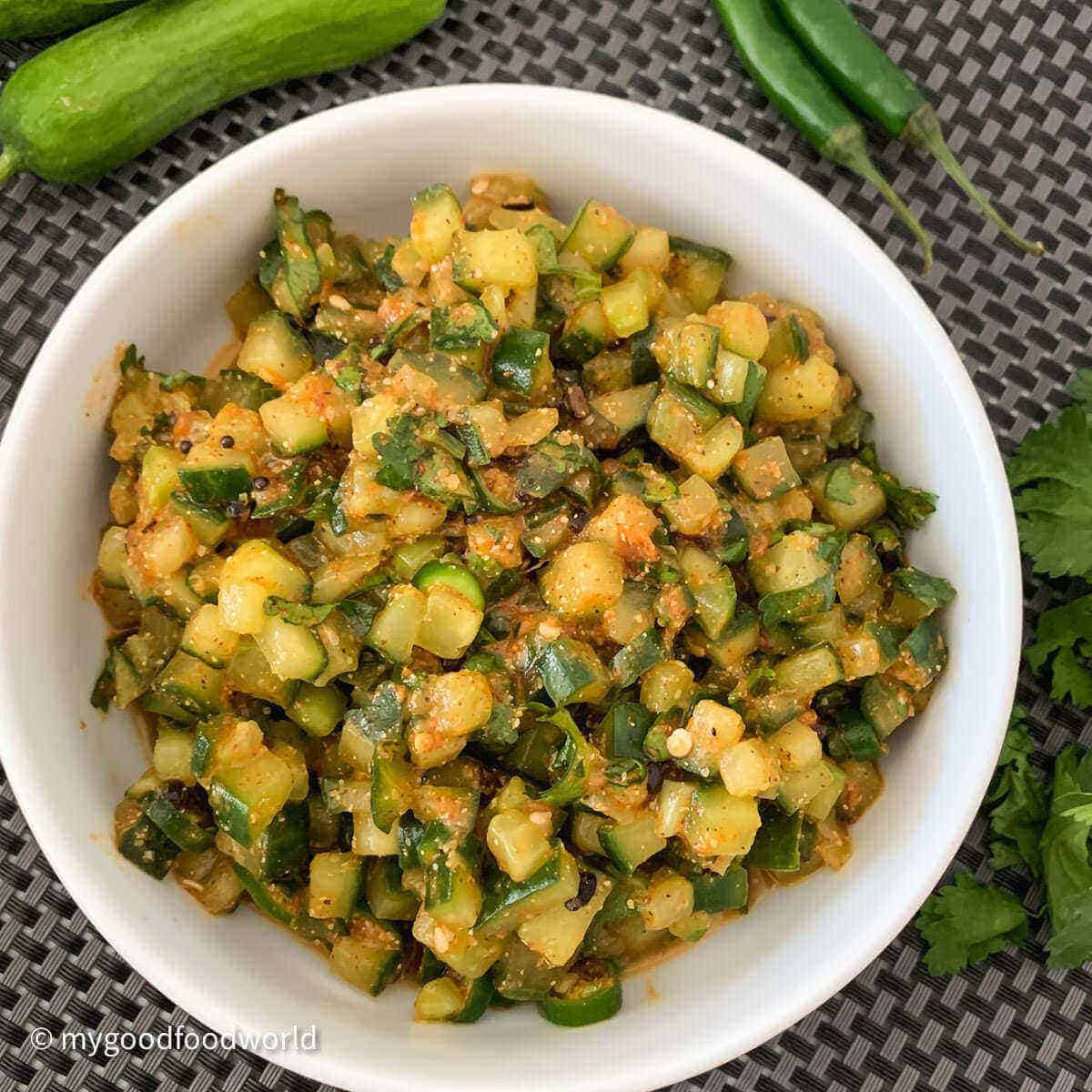 Spicy cucumber salad placed in a white round bowl on a black mat with fresh kheera, green chillies and fresh cilantro placed on the side of the bowl.
