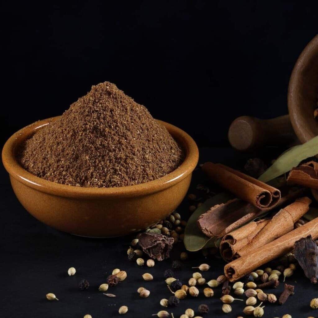 Powdered garam masala is placed in a round brown colored bowl and some whole garam masala spices are placed next to the bowl against a black backdrop.