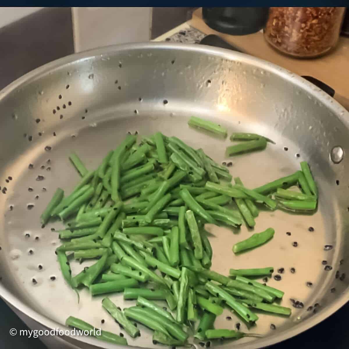 Tender green beans being sauteed in oil and nigella seeds in a steel frying pan.