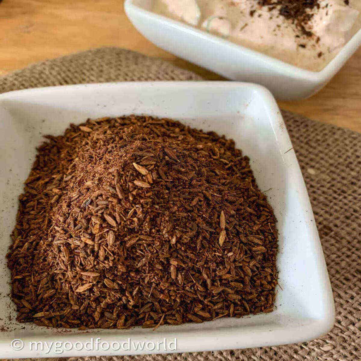 Roasted powdered cumin placed in a white bowl and placed on a burlap. next to the bowk is another bowl with yogurt dip and bhuna jeera topping.