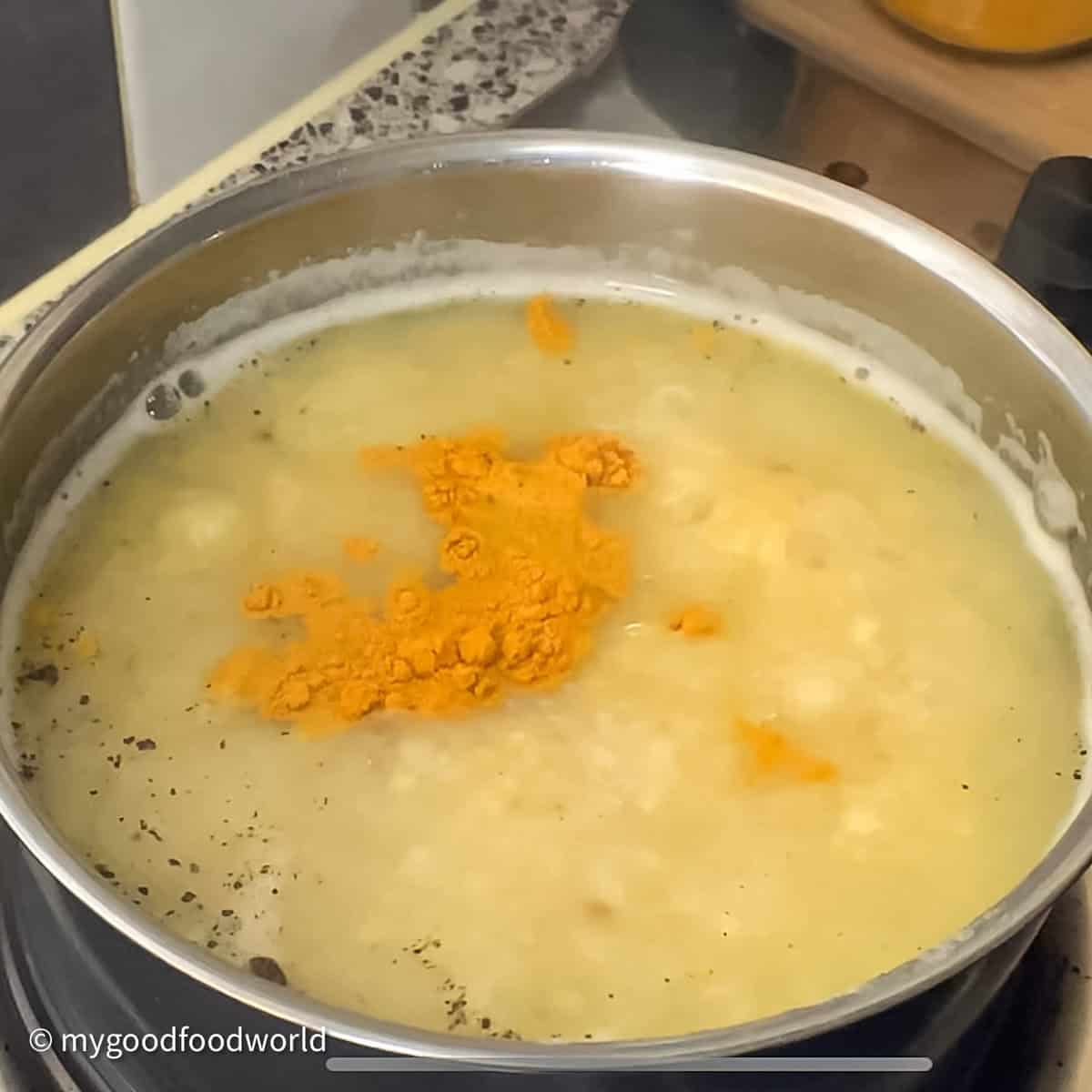 A pot of yellow soup with orange seasoning on a black stovetop. A yellow spatula or spoon is on the right. 