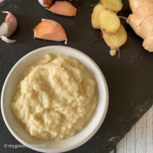Homemade ginger garlic paste placed in a white round bowl which is placed on a black board and with garlic cloves and ginger pieces on the side.