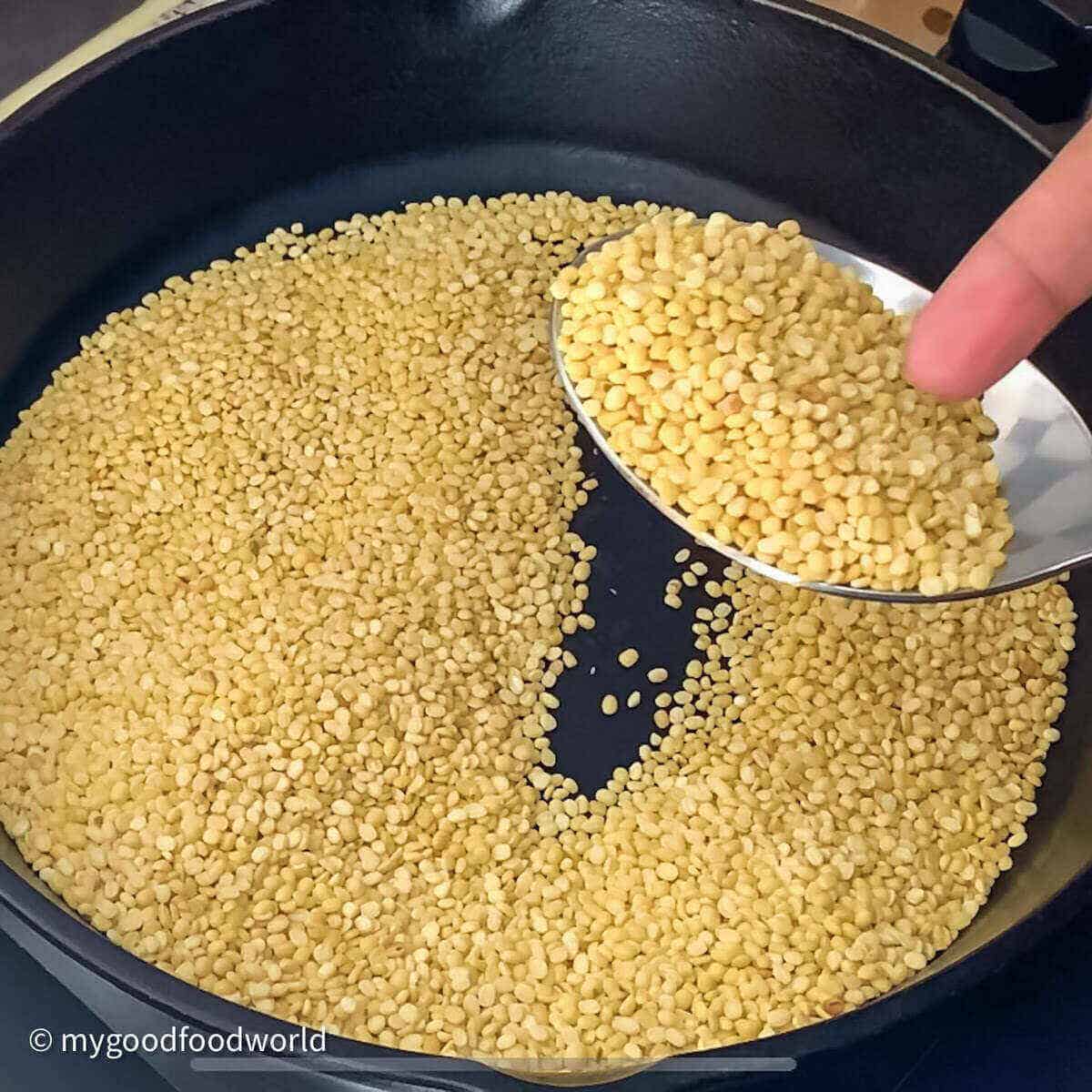 Yellow lentils roasting in a black frying pan with a silver spoon held by a finger.