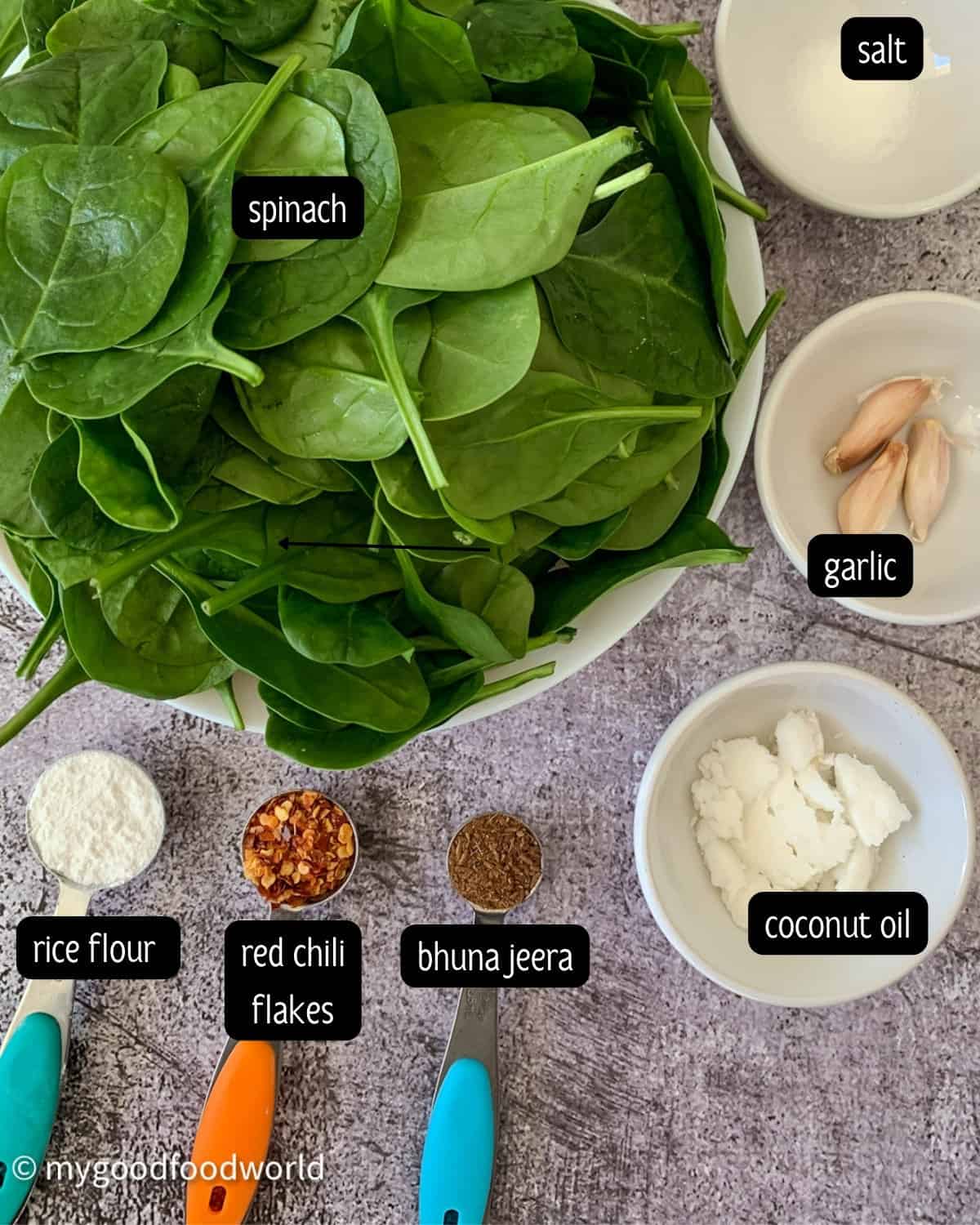 Ingredients for spinach stir fry placed in small white bowls and measuring spoons which are placed on a light grey background.