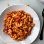 Vegetarian macaroni and tomatoes dish served in a round white bowl-plate which is placed on a light grey mat.