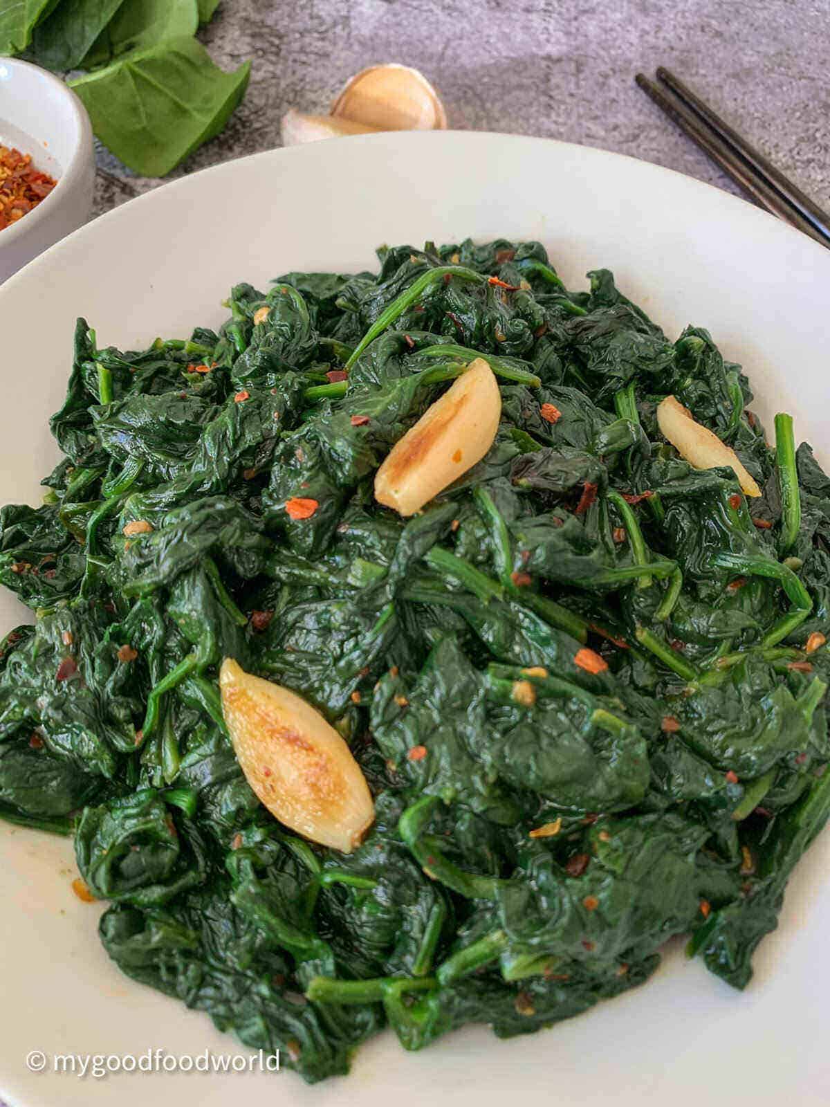 Stir fried spinach with whole garlic placed in a white bowl plate with chili flakes, chopsticks and fresh spinach leaves on the side.