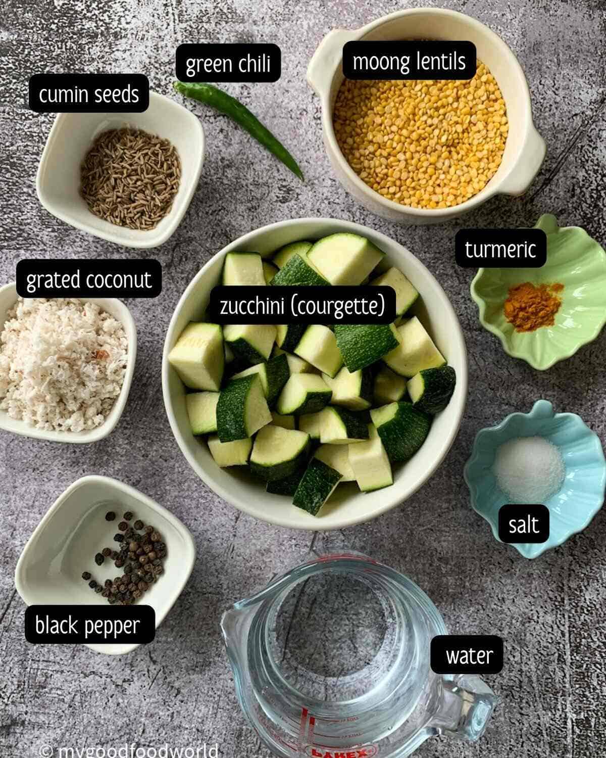 Ingredients for the zucchini kootu recipe are placed in small bowls and arranged on a dark grey and white patterned background.