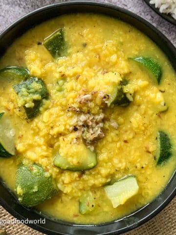 Zucchini and mung lentils kootu served in a round black color bowl. The bowl is placed on a piece of burlap and has a spoon and a plate of rice placed next to it.