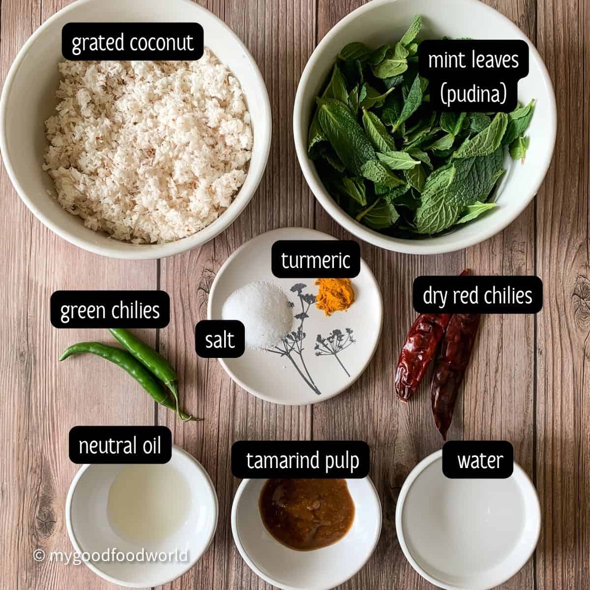 Mint leaves, grated coconut, oil, tamarind pulp, water, two green chilies, and two dry red chilies placed in white bowls and arranged on a brown textured background.