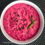 Beet raita served in a round white bowl with roasted cumin, green chilies, and fresh herbs as garnish.