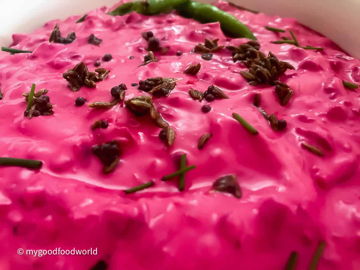 A close-up photo of bright pink beet raitha with tadka of roasted cumin, chilies, and fresh herbs.