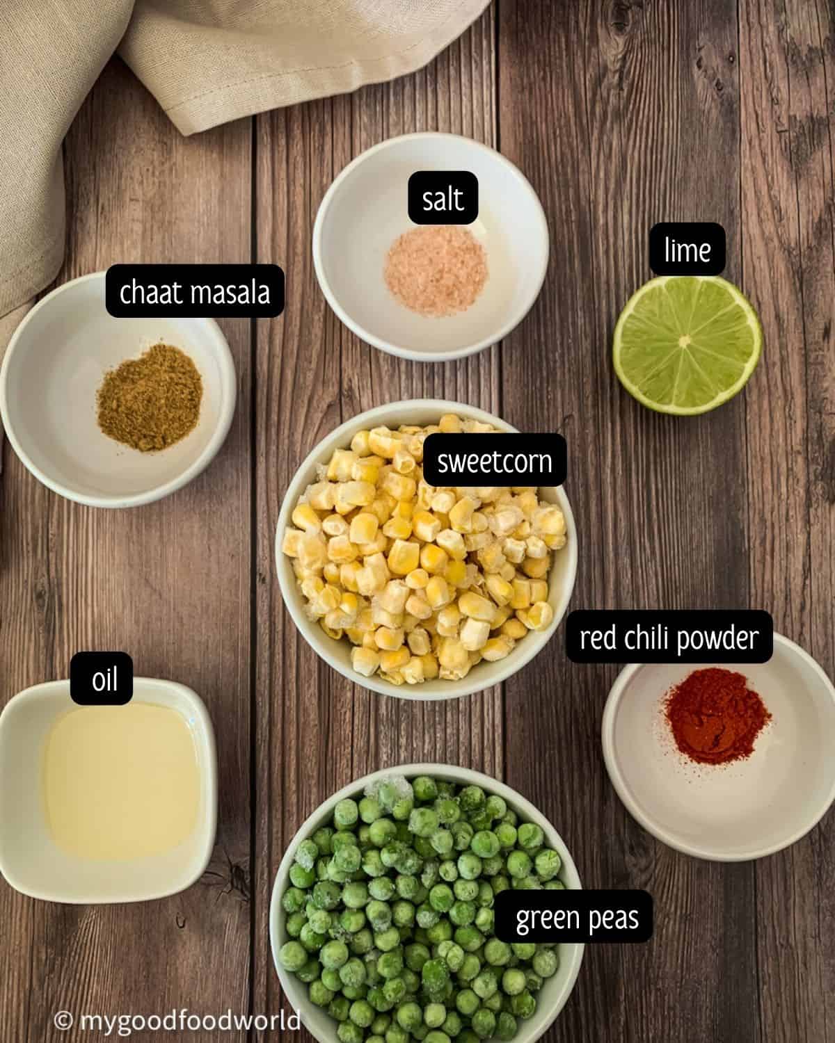 Ingredients for peas and corn salad are placed in white bowls which are placed on a dark brown textured backdrop.