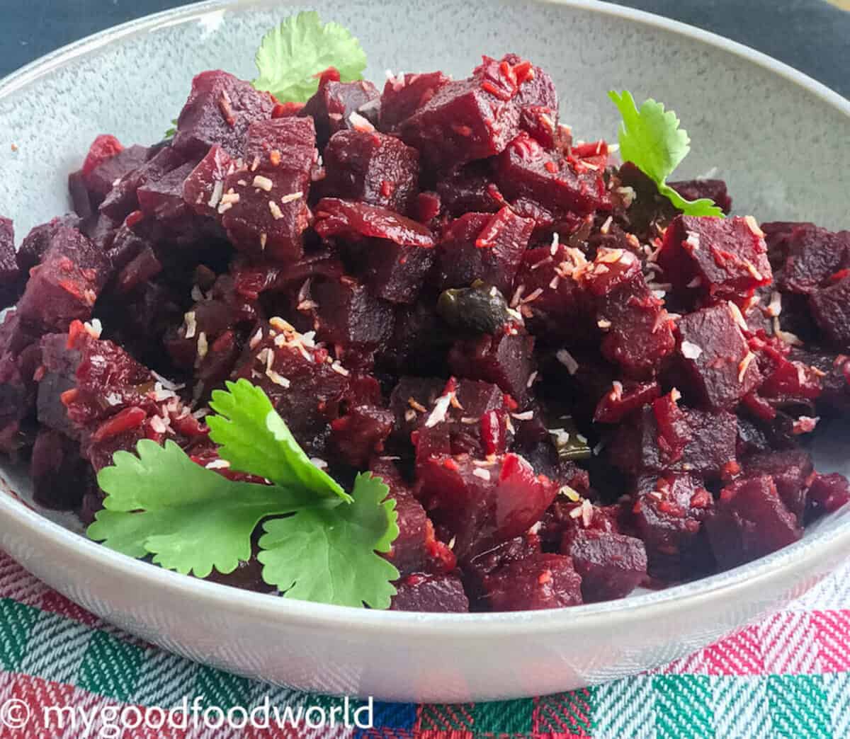 Soft cooked beetroot sautéed with spices and a garnishing of coconut flakes is placed in a round bowl which is placed on a colorful cloth.
