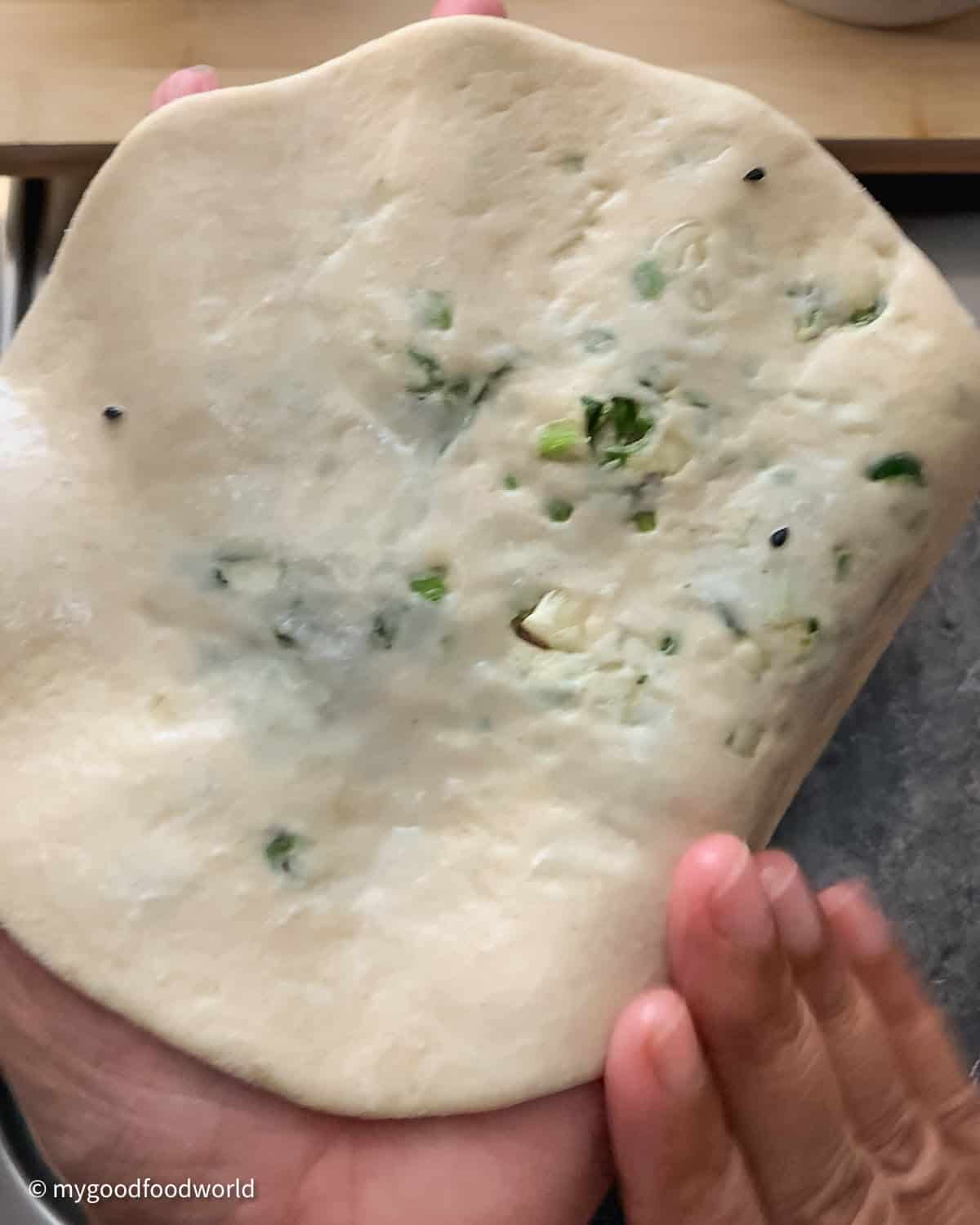 Water being applied on one side of a rolled out naan bread. The naan has specks of green on it.