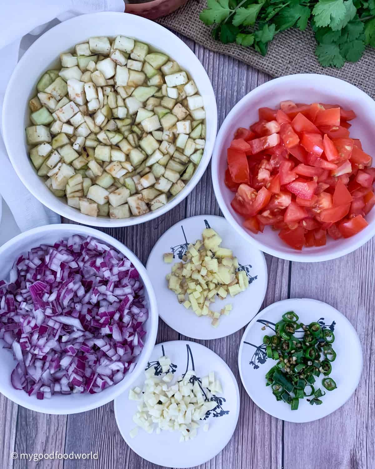 Finely chopped eggplants, onion, ginger, garlic, chili pepper, and tomatoes placed in round bowls and plates on a brown table.