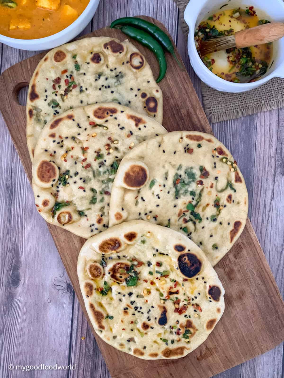 Naan breads with spicy toppings such as nigella seeds, cilantro, and red pepper flakes are placed on a wooden board overlapping each other. Next to the board, there is some ghee with spices in a round, white bowl, two green chilies, and a bowl of curry.