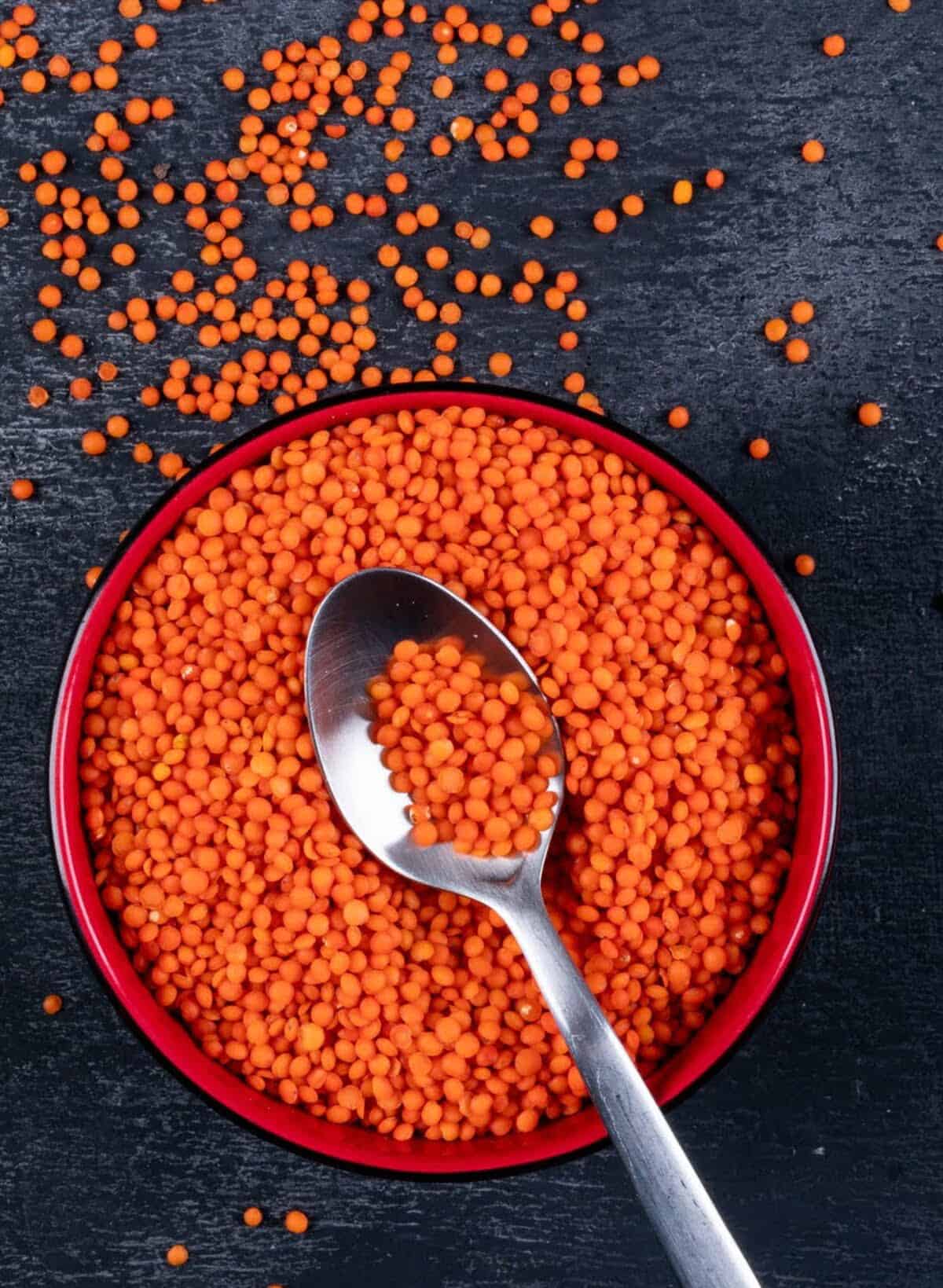 Sabut masoor dal are placed in a round red bowl with a metal spoon scooping some of the lentils from the bowl. The bowl is placed on a dark backdrop.