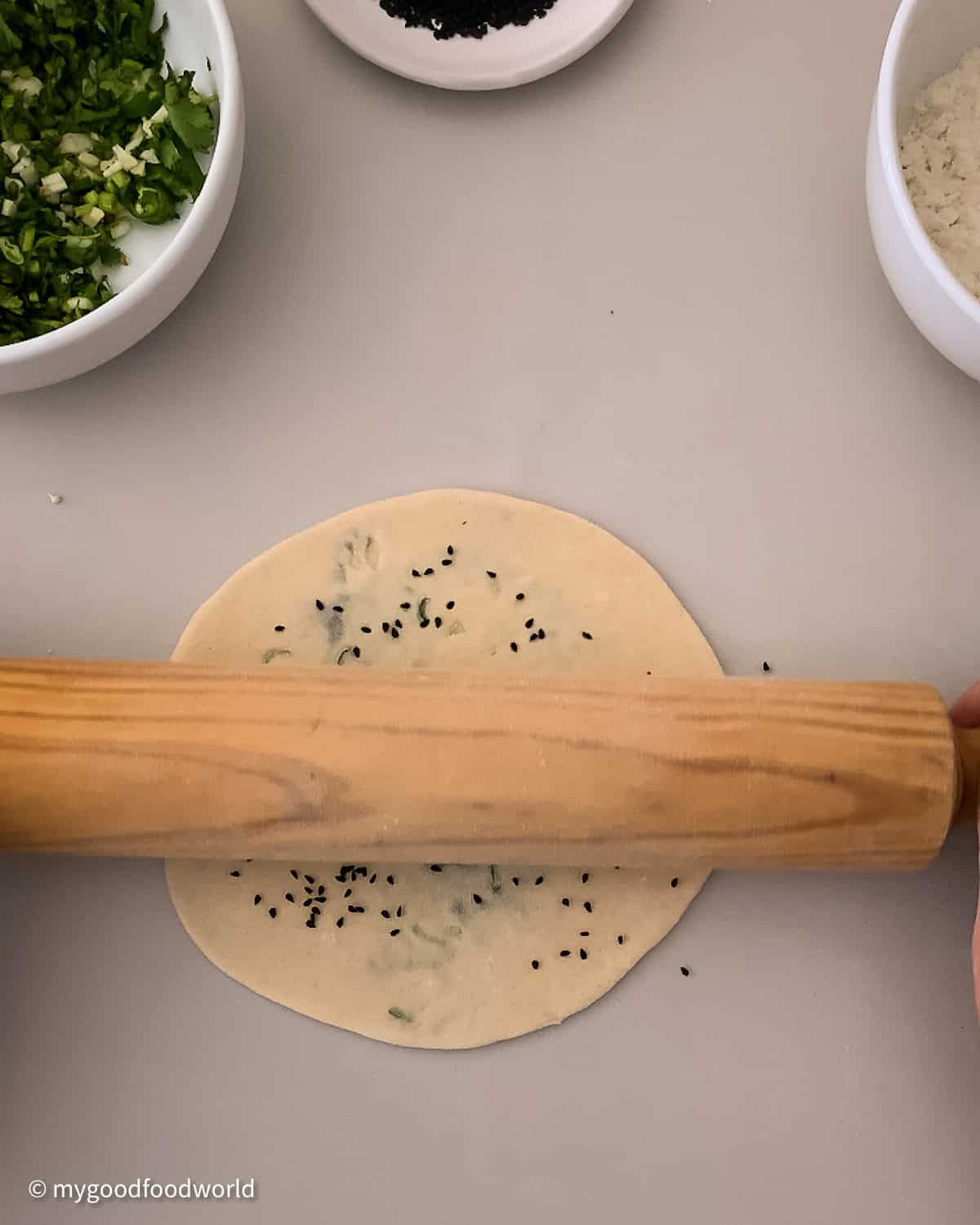Nigella seeds that are sprinkled on a flatbread are being pressed down into the naan with a rolling pin.