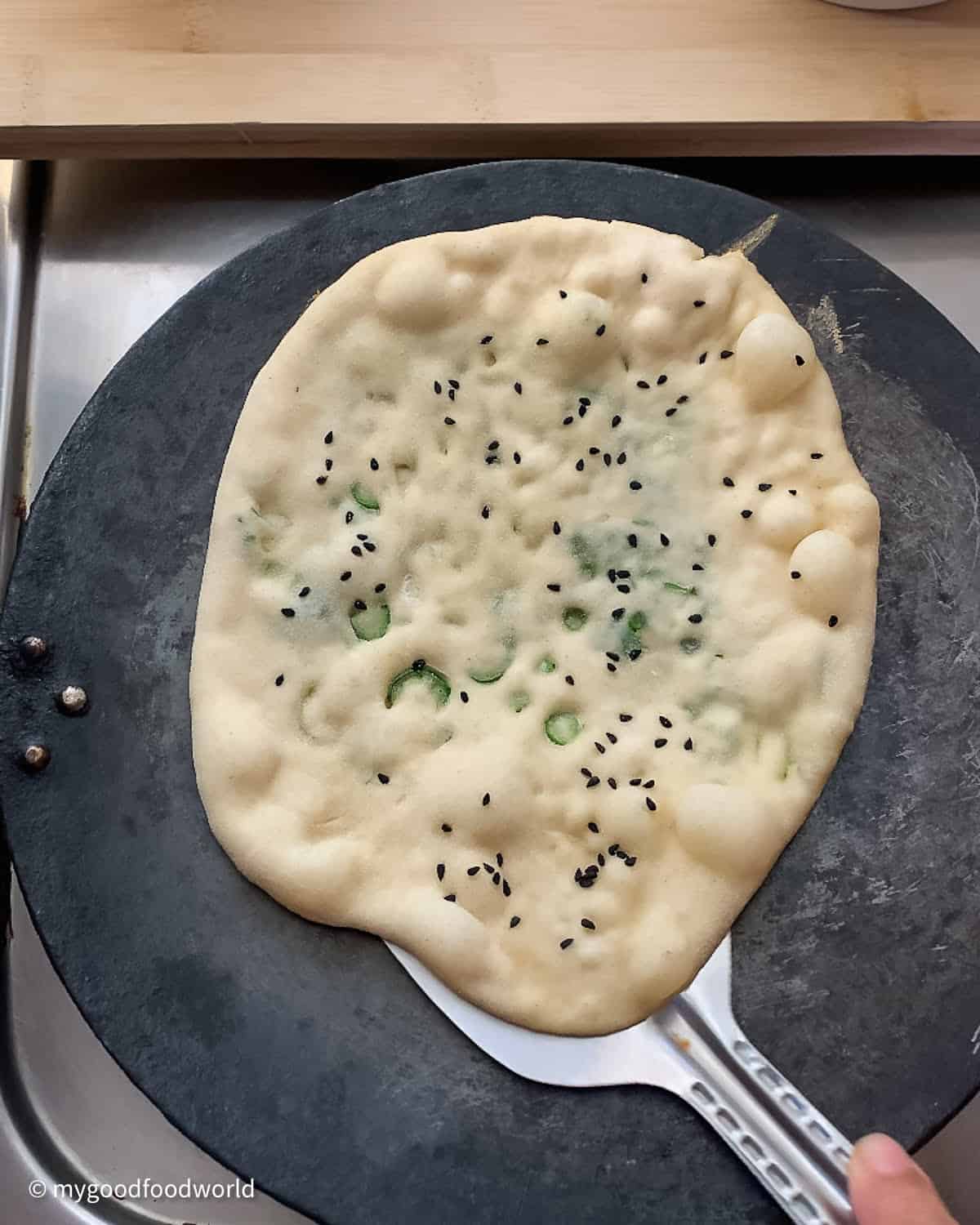 A flatbread is being roasted on a cast iron roasting pan. The bread has bubbles on its surface and a metal spatula is being slid under the bread.