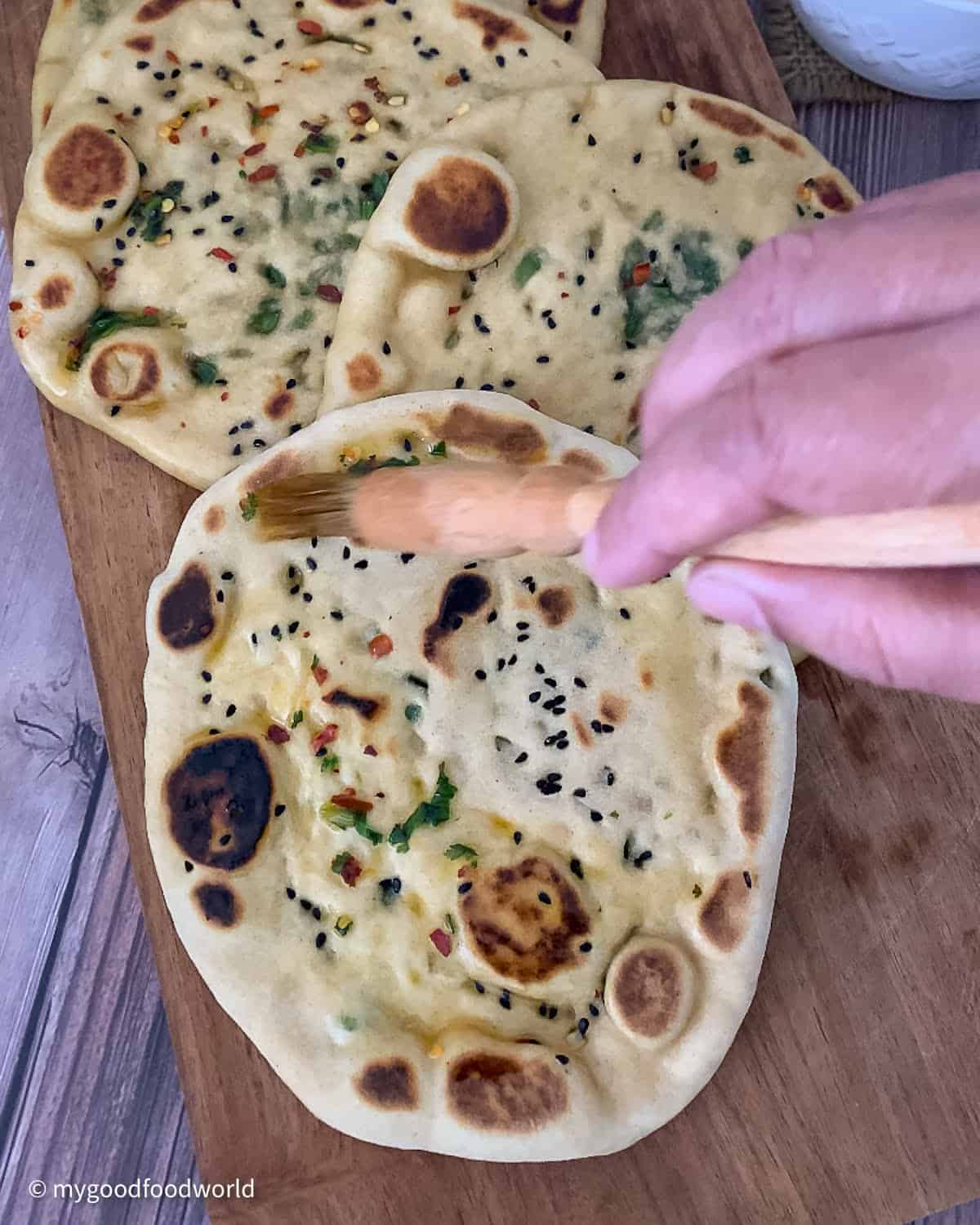 Ghee that has fresh herbs and red pepper flakes in it is being applied on a well roasted naan bread with a wooden brush.