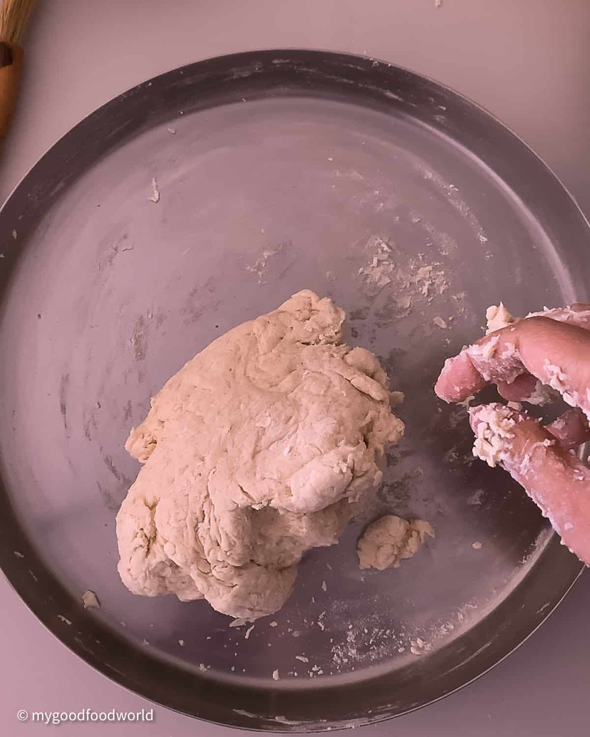 Plain flour sticky dough is kept on a steel plate. A hand is seen with some dough on it. 