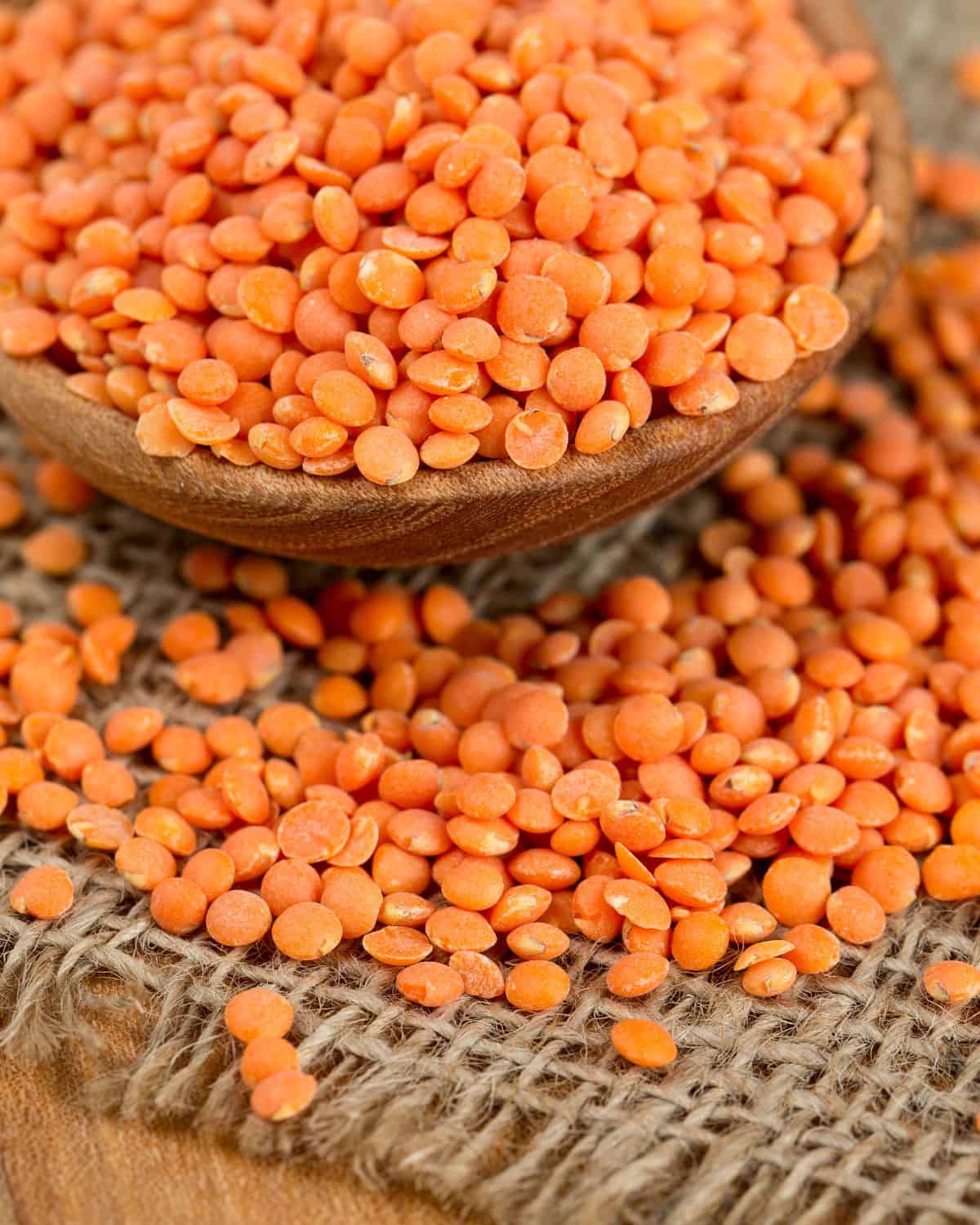 Whole red lentils are placed in a wooden bowl. The bowl is placed on a piece of burlap. Some of the lentils have spilled on the burlap.
