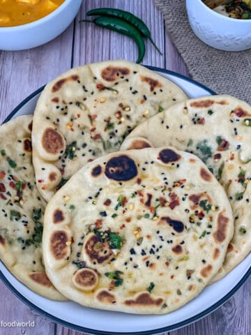 Four small-sized naans studded with red pepper flakes, nigella seeds, cilantro, and ghee, placed in a white round bowl. The bowl is placed on a textured brown background.