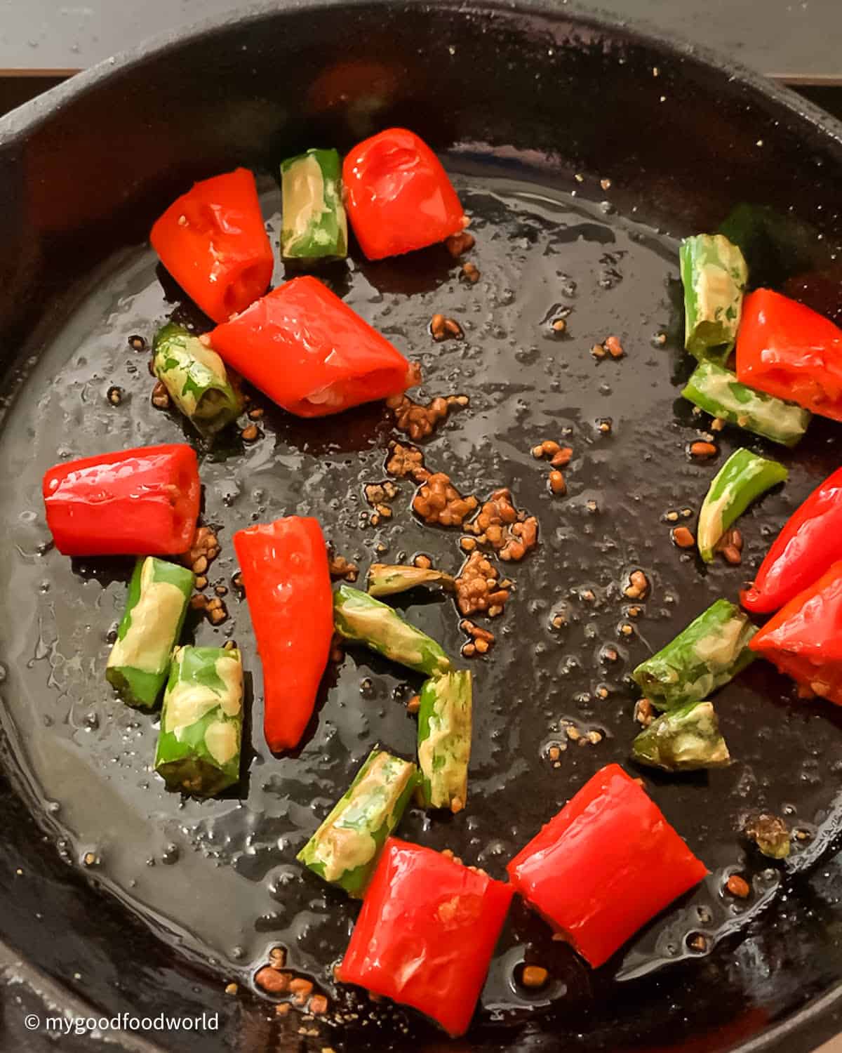 Fresh red and green chilies are frying in some oil and with whole spices in a cast iron pan.