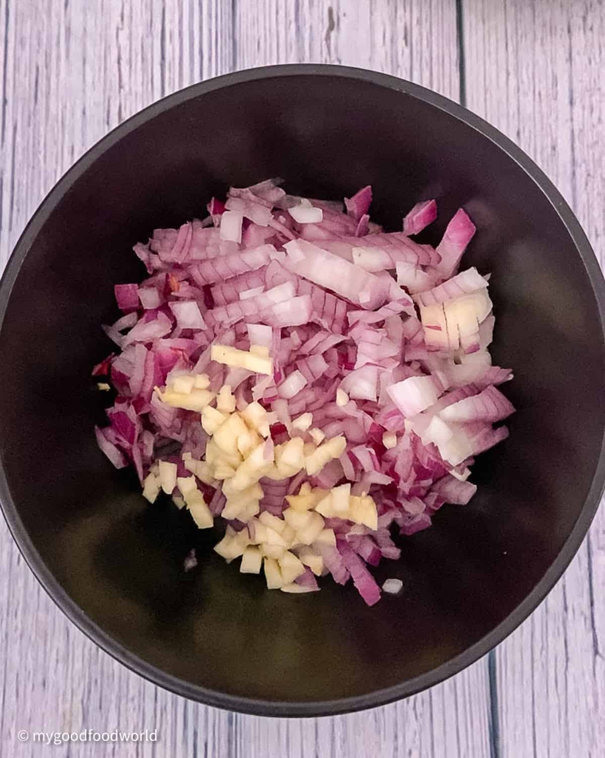 Finely chopped onion and garlic for paneer spinach curry are placed in a round black bowl. The bowl is placed on a light blue patterned background.