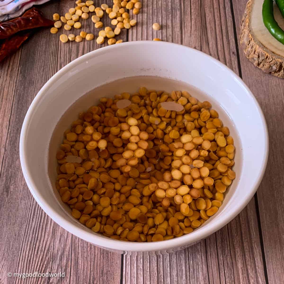 Roasted chickpea lentils are soaking in water in a round white bowl. The bowl is placed on a patterned brown backdrop with some unroasted chana dal and chilies placed on the backdrop.