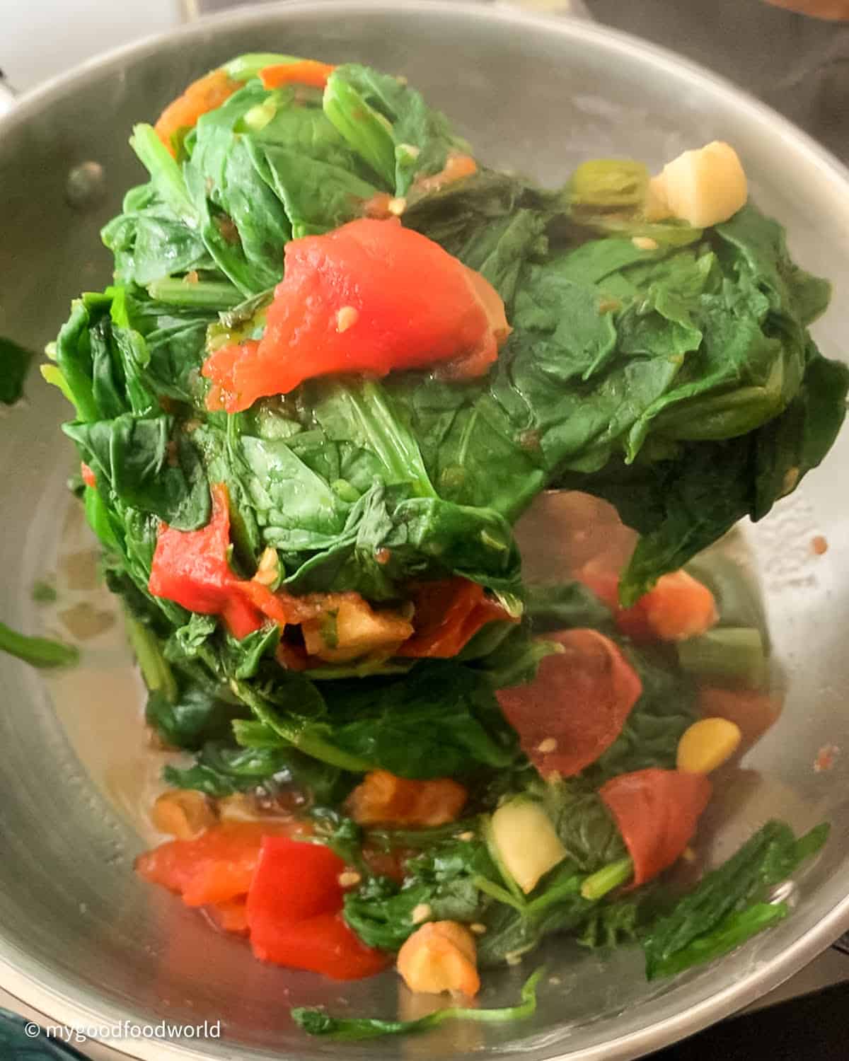 For Punjabi palak paneer sabji, spinach is being cooked with tomatoes, garlic, and ginger being scooped up with a spoon from a stainless steel wok.