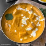 Mughlai shahi paneer is topped with some fresh cream and cilantro is served in a round steel bowl. The bowl is placed on a piece of burlap.