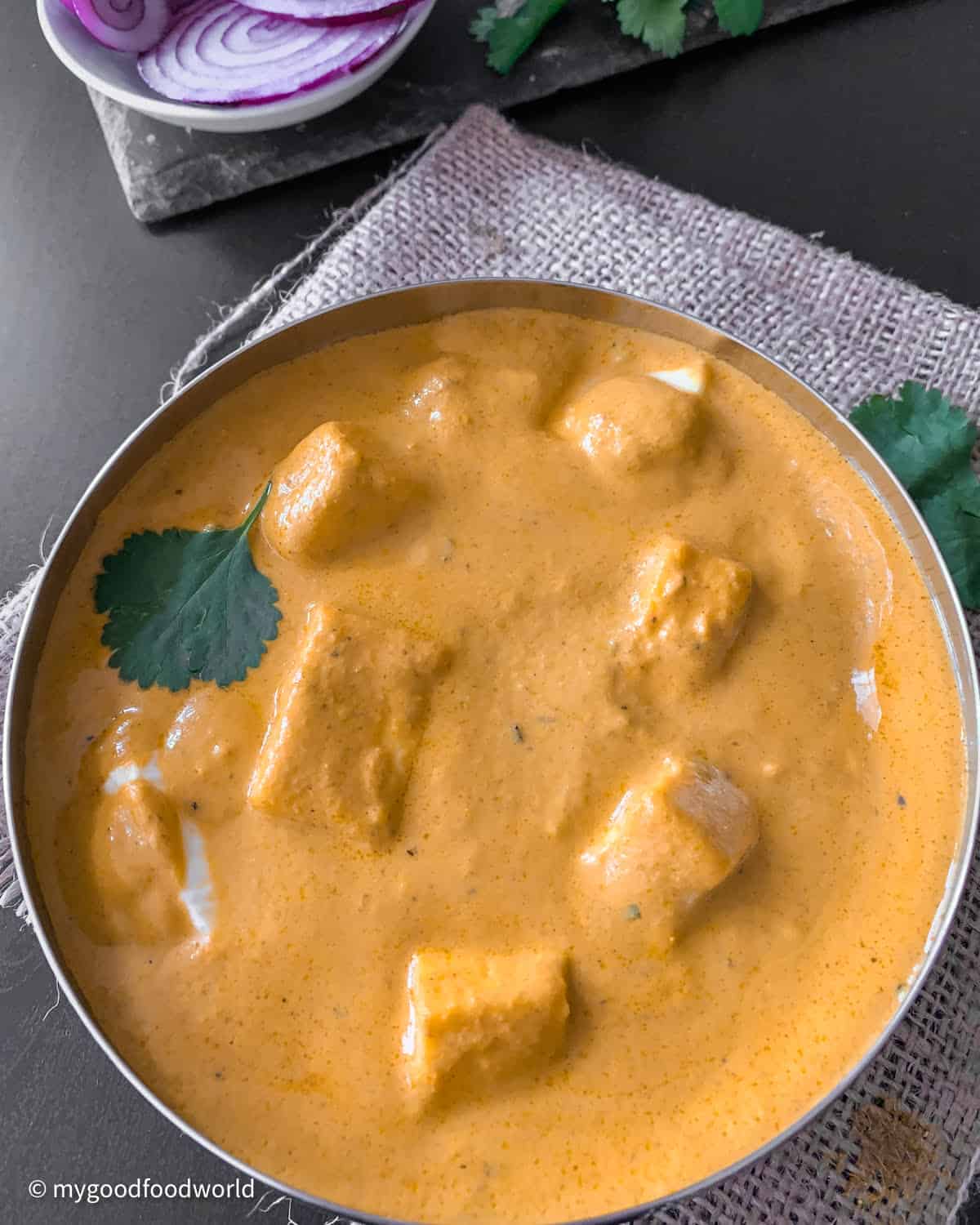 A photo of a creamy orange gravy with paneer chunks in a white bowl on a gray countertop. There is a red onion and cilantro next to the bowl.