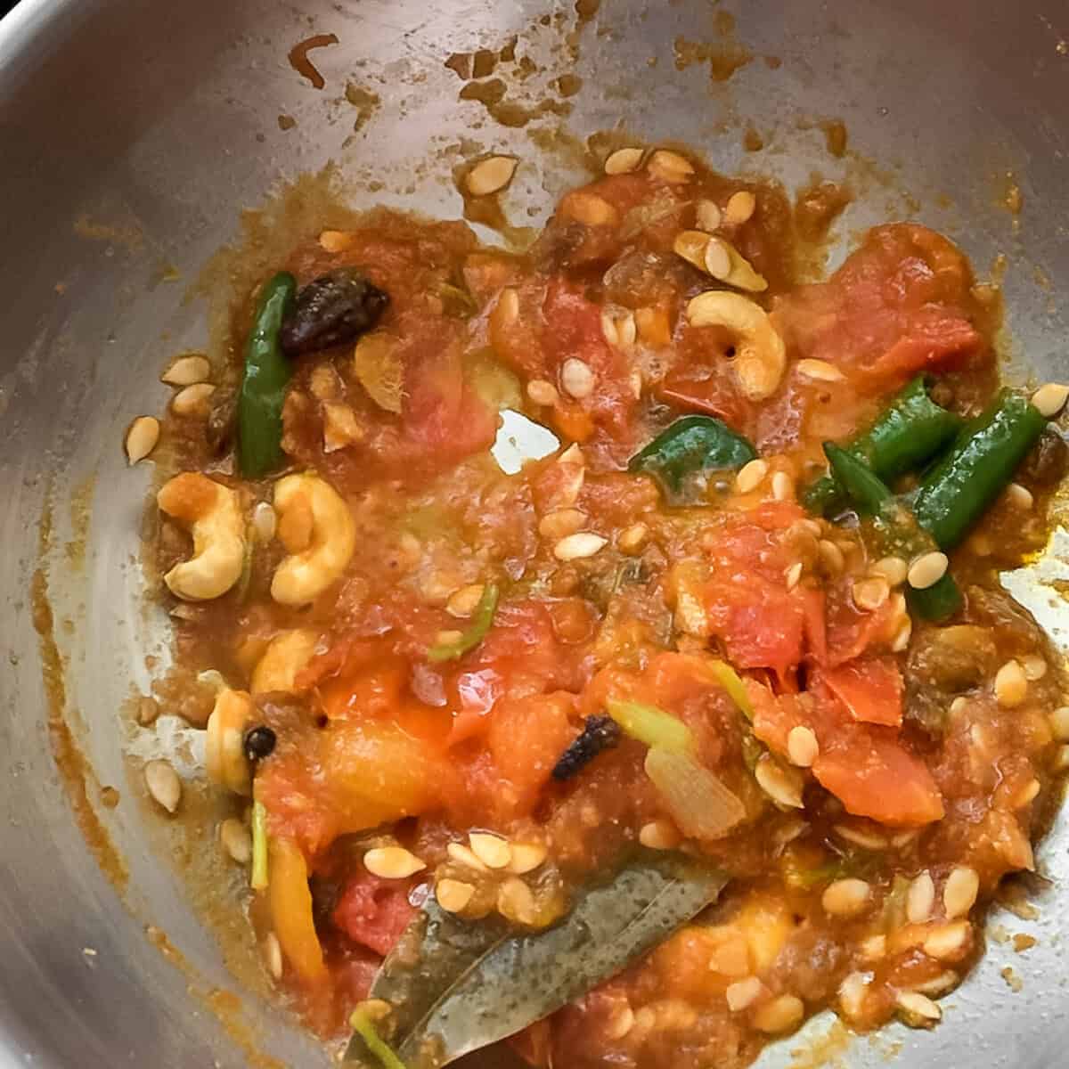 A mixture of tomatoes, melon seeds, cashew nuts, bay leaf, and whole spices cooking in a stainless steel wok to make a shahi sauce.
