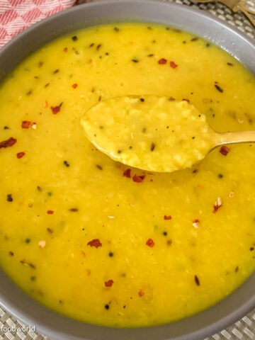 A bowl of yellow lentil soup with a gold spoon on a red and white checkered tablecloth. The soup has black and red specks.