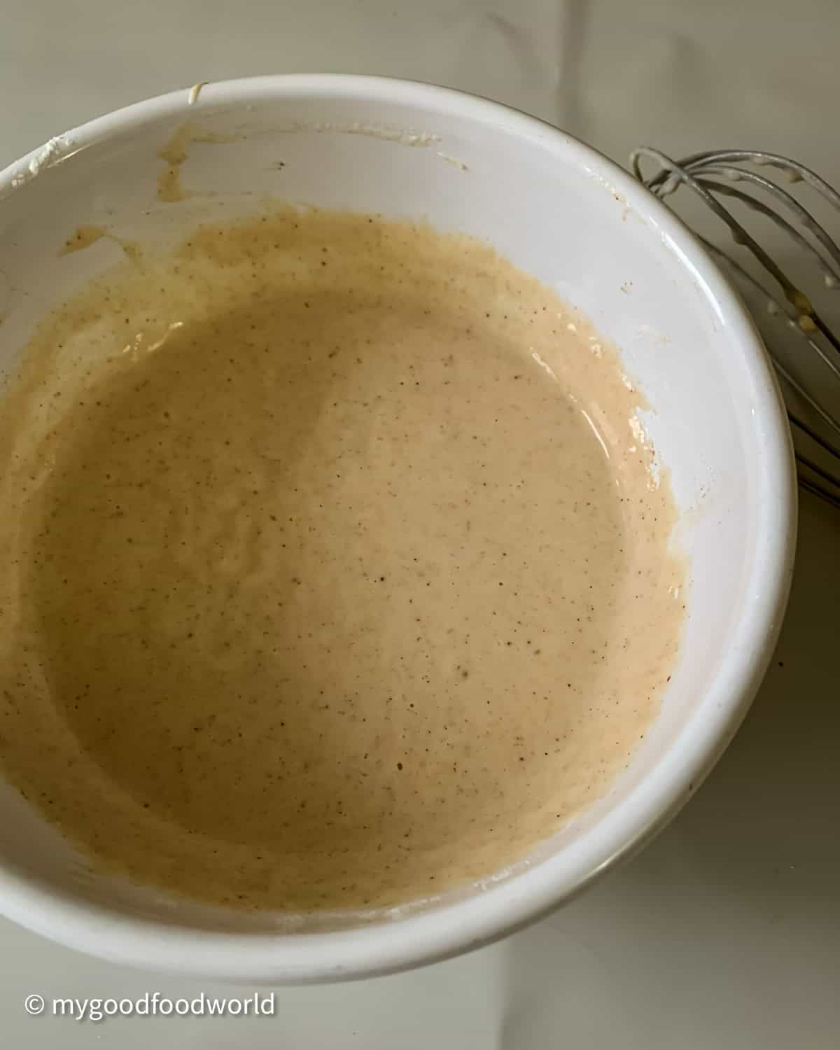 Batter for pancake is placed in a white round bowl. The batter has brown specks in it which looks like cinnamon powder.