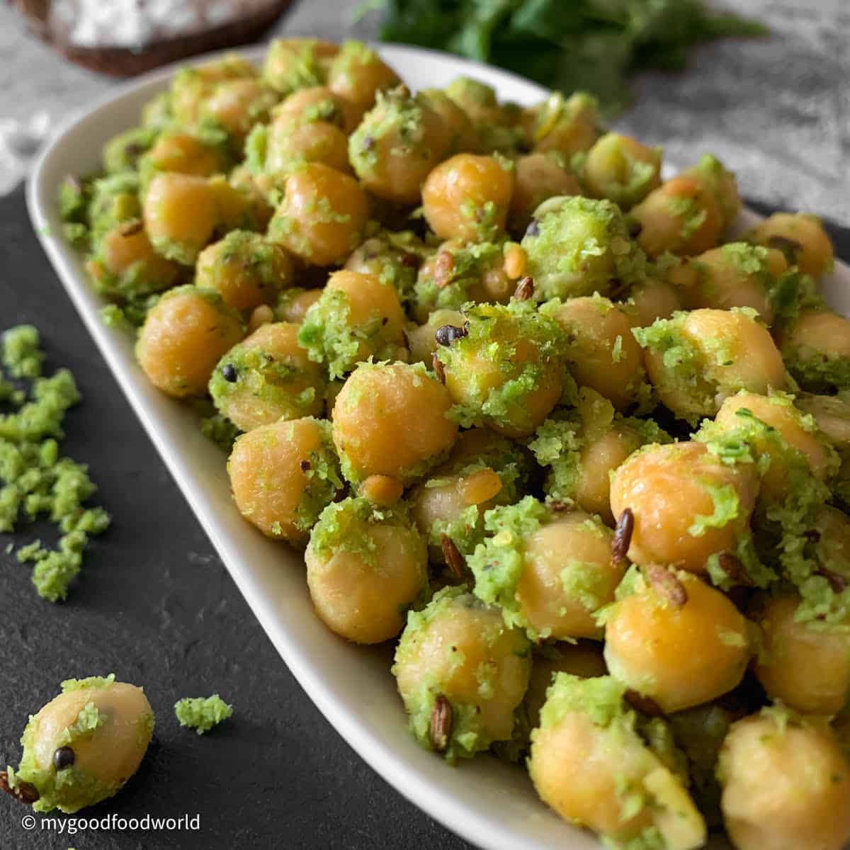 Cooked whole chickpeas salad with garnish of fried cumin, mustard seeds and white lentils in a green, coconut paste is placed in a white bowl-plate.