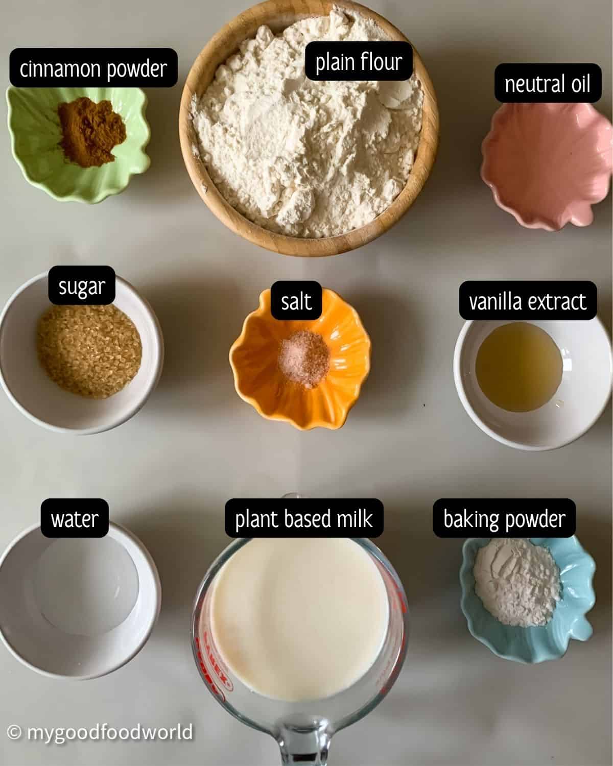Ingredients for vegan pancake recipe without eggs, namely, all purpose flour, plant based milk, baking powder, vanilla extract, sugar, cinnamon powder, oil, and water are placed in small bowls next to each other.