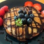 One fluffy eggless pancake is placed on a bluish green plate. There is a crisscross pattern made on the pancake with chocolate sauce. Some fresh blueberries and strawberries are placed on the pancake and next to it.