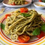 Spaghetti with fresh basil, roasted tomatoes, and pesto sauce with a garnish of nutritional yeast flakes is placed in a round, colorful plate.