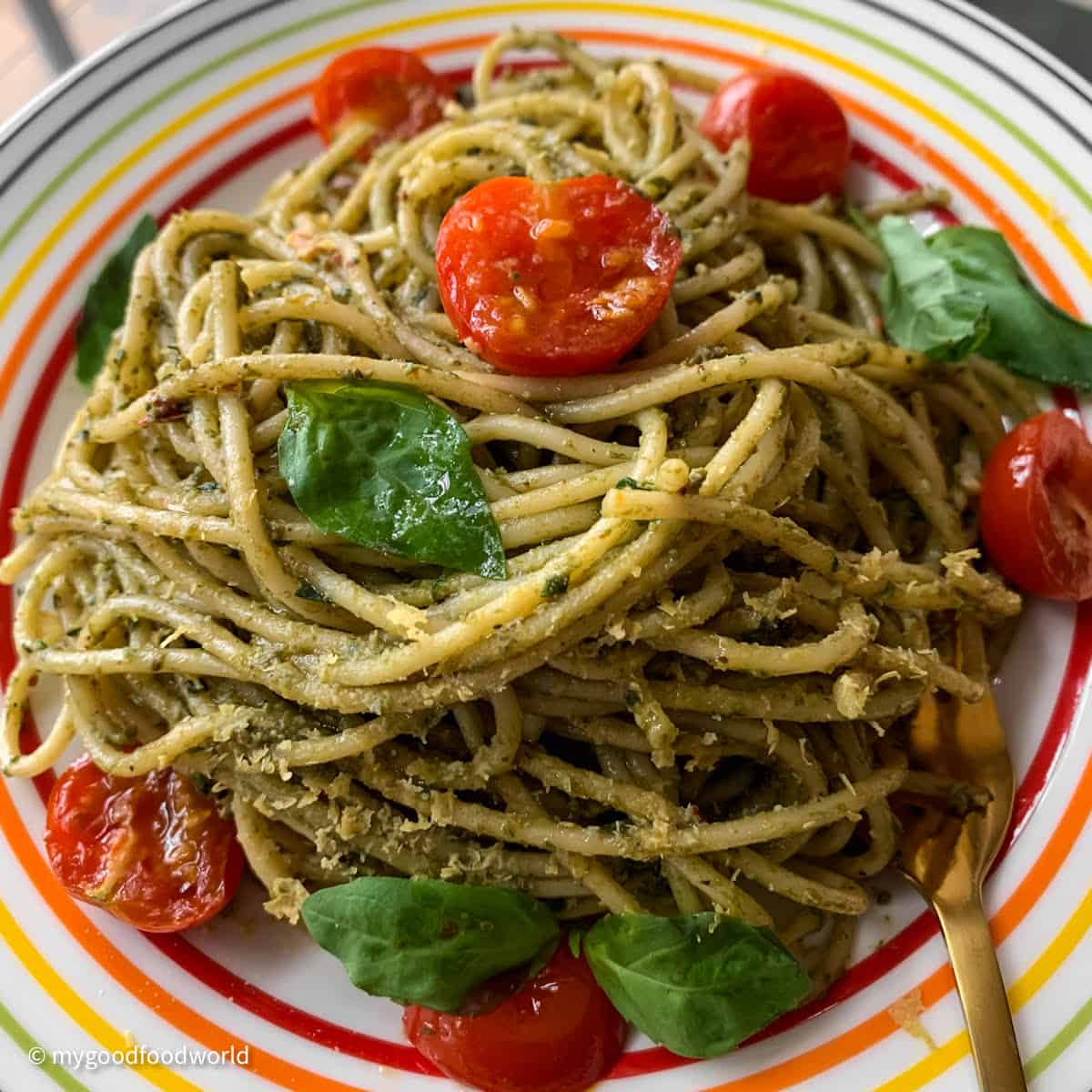 Cooked pasta that has been tossed in pesto sauce is placed in a mounds on a colourful plate. The pasta is topped with roasted cherry tomatoes and yeast flakes. Some fresh basil leaves are placed on the dish.